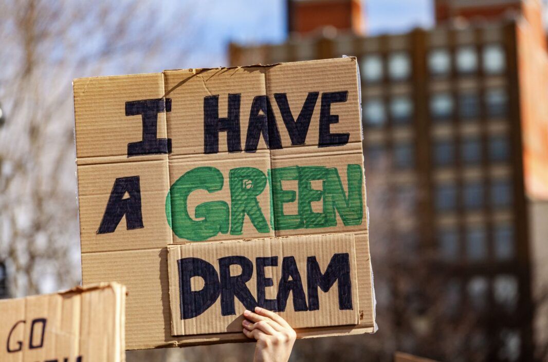 A close-up of a hand holding up a cardboard sign with the words "I HAVE A GREEN DREAM" written on it. The letters "I HAVE A" are written in black, while the word "GREEN" is emphasized in green color. The word "DREAM" is again in black. The background is blurred but appears to be an outdoor setting with a building visible, suggesting that this may be from a protest or rally for environmental causes.